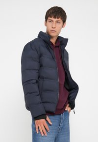 Marineblå pufferjakke med høy krage, glidelås og quiltet tekstur. Brukt over en burgunderrød hettegenser og blå jeans.