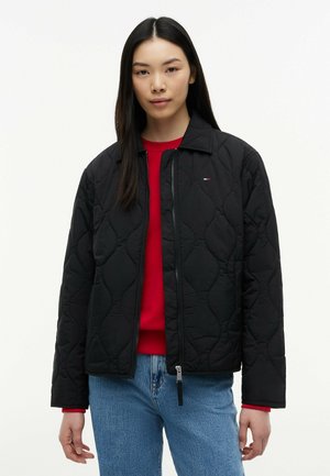 Kobieta ubrana w czarną pikowaną kurtkę na czerwonym topie i niebieskich dżinsach, stojąca na tle jednolitego białego tła.