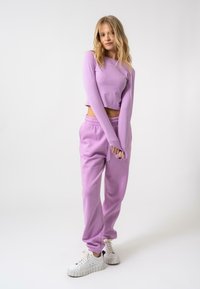 Top corto a maniche lunghe di un leggero viola abbinato a pantaloni jogger coordinati, dotati di cintura elastica e polsini a costine, indossato con sneakers bianche.
