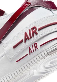 Biała tenisówka z czerwonymi akcentami, wykonana z teksturowanego materiału, z wyraźnym logo „AIR” i gumową podeszwą z wzorzystym bieżnikiem.