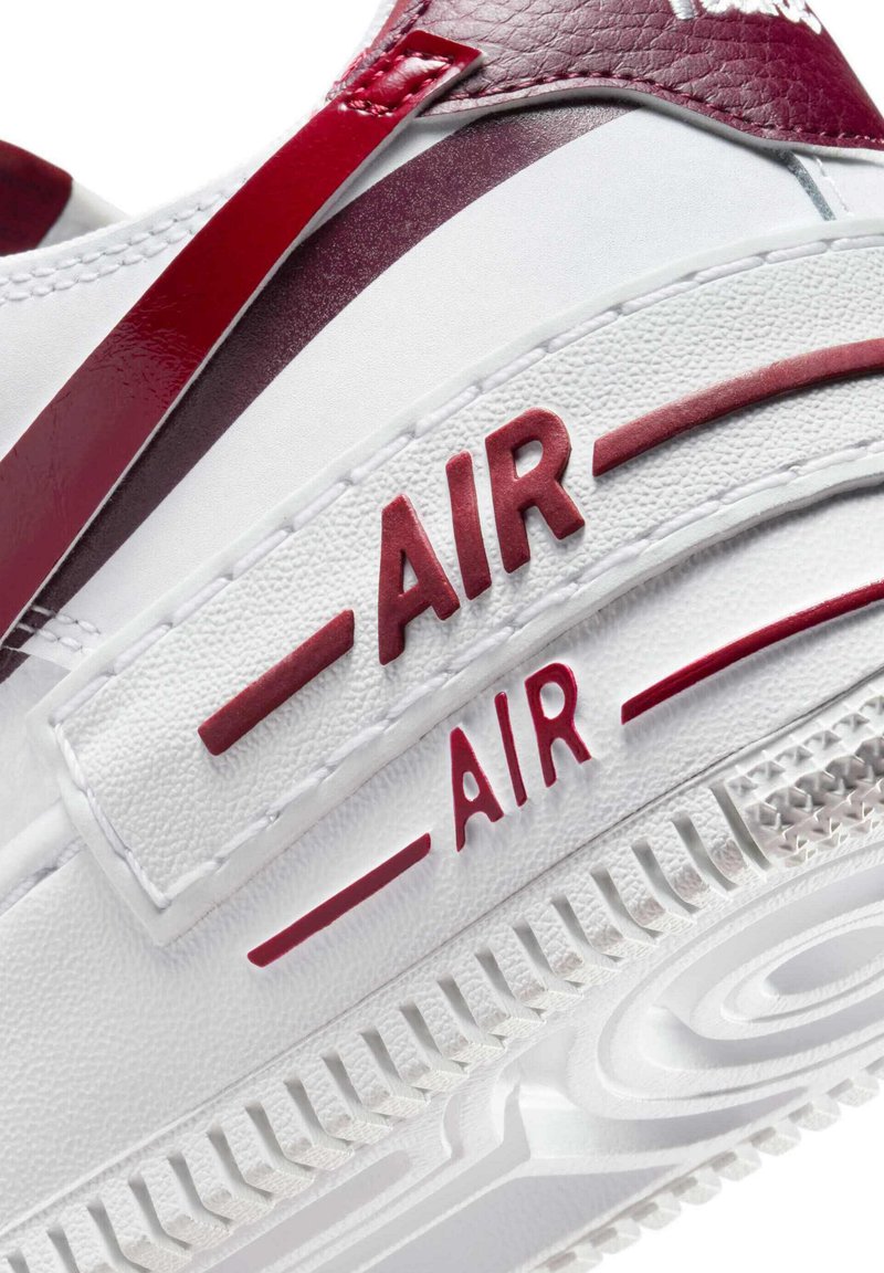 Hvid sneaker med røde detaljer, der har tekstureret materiale, tydelig "AIR" branding og en gummiydersål med mønstret slidbane.