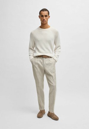 Maglione a coste bianco, pantaloni a quadri beige, cintura in pelle marrone e scarpe abbinate. Il modello sta in piedi con le mani in tasca su uno sfondo neutro.