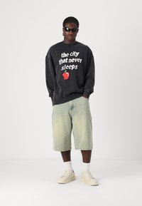 Black sweatshirt with graphic text and red apple design, paired with light green shorts. Model wears sunglasses and white sneakers.