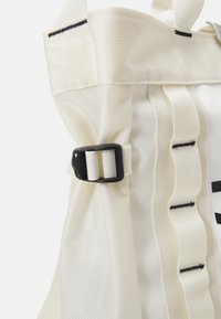 Sac à dos en tissu blanc avec un matériau texturé, une boucle noire réglable et des accents noirs verticaux le long des bretelles.