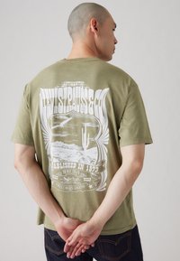 Olive green cotton t-shirt featuring a large white graphic print of a desert scene with cacti and birds, and vintage-style text on the back.