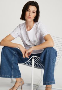 Biała koszulka z krótkim rękawem z nadrukowanym tekstem i akcentami, zestawiona z szerokimi niebieskimi dżinsami. Model siedzi na białym, drucianym krześle.