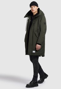 Jonge man in een donkergroen capuchonparka, zwarte beanie, zwarte hoodie, donkere jeans en zwarte chunky boots, staand tegen een effen achtergrond.