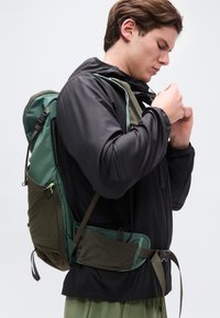 Νεαρός άνδρας που ρυθμίζει τον ιμάντα στο στήθος ενός πράσινου σακιδίου πεζοπορίας, φορώντας μαύρο μπουφάν και πράσινο παντελόνι, απομονωμένος σε λευκό φόντο.