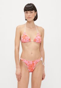 Bikini rose léger avec motif floral, comportant des liens blancs. Haut triangle et bas taille basse. Texture de tissu douce.