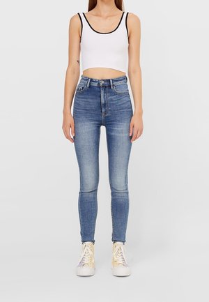 Woman wearing high-waist blue skinny jeans, white crop tank top with black trim, and cream high-top sneakers, standing against plain background.