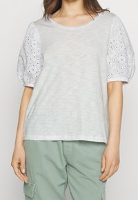 Světle šedé tričko s jemnou texturou a kulatým výstřihem, které má bílé vyšívané nabírané rukávy. Nosené s světle zelenými kalhotami.