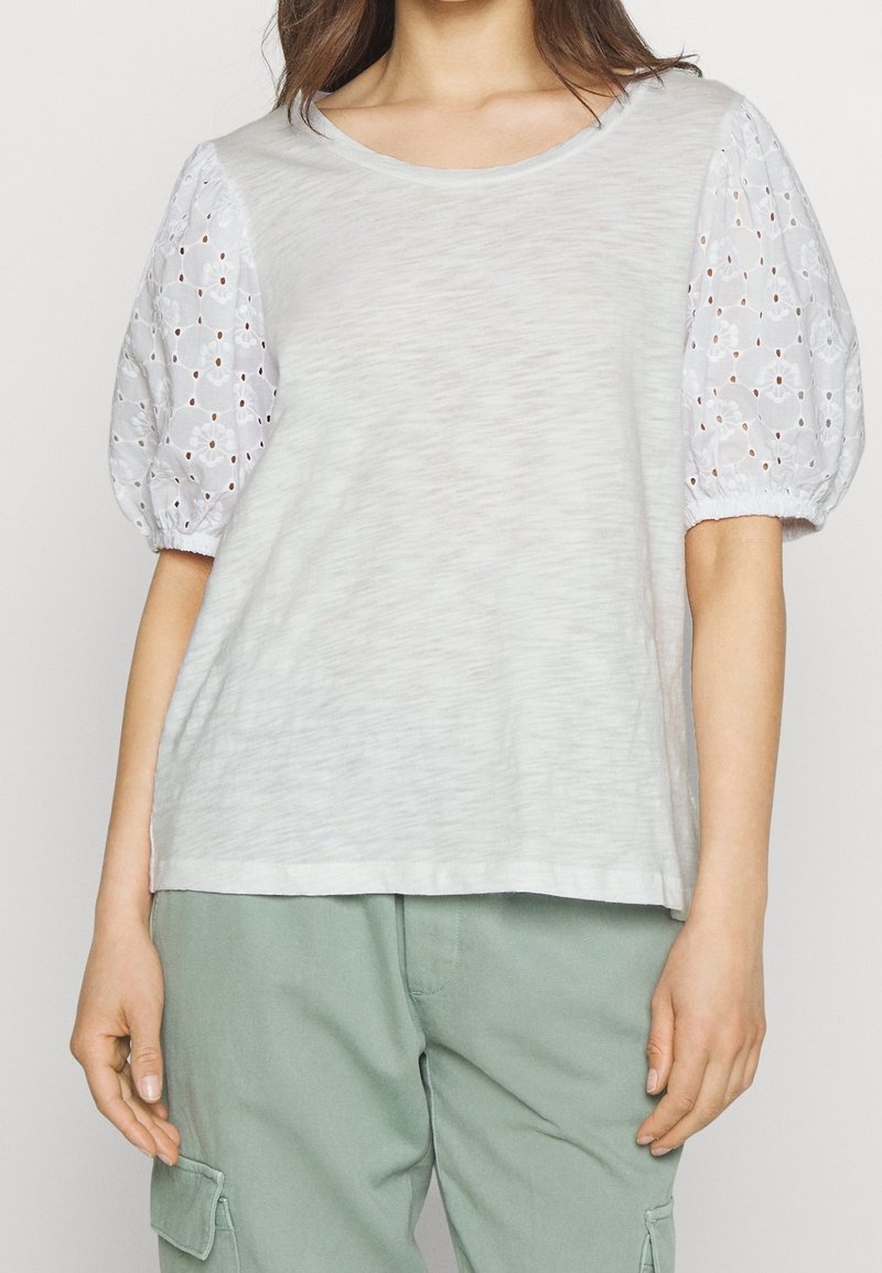 Světle šedé tričko s jemnou texturou a kulatým výstřihem, které má bílé vyšívané nabírané rukávy. Nosené s světle zelenými kalhotami.