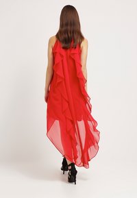 Robe en chiffon rouge avec des volants superposés le long du dos et un ourlet asymétrique. Portée avec des sandales à talons hauts noires.