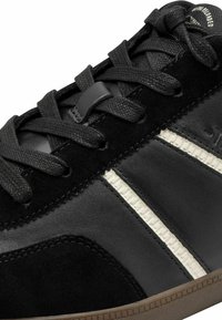 Närbild av svart sneakers med mocka- och läderpaneler, svarta snören, vita sidremsor och brunt texturerat sula.