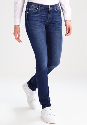 Persoon die slank vallende donkerblauwe spijkerbroek, witte sneakers en een wit shirt met lange mouwen draagt, met gekruiste benen staand op een witte achtergrond.