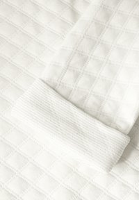 Weißer gestepptes Gewebe mit einem Diamantmuster, mit einem gerippten Ärmelsaum in einer glatten weißen Textur, das die Nähdetails hervorhebt.