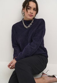Paarse, fuzzy trui met hoge hals, gecombineerd met een zilveren chunky ketting en oorbellen. Zwarte broek en zilveren heeled sandalen.