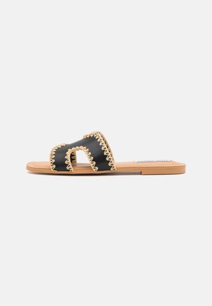 Black slide sandals featuring a cut-out design, beige scalloped edging, flat tan sole, and minimalistic style.