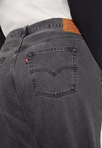 Jean en denim gris avec un design classique à cinq poches, poches arrière cousues avec un motif en "V" incurvé, et une étiquette en cuir à la taille.