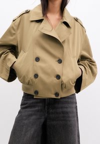 Kvinde iført en cropped beige dobbeltbrystet jakke med sorte knapper og mørke højtaljede jeans, hænderne i lommerne.