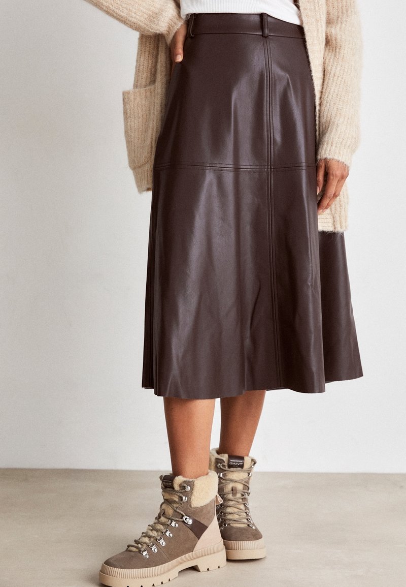 Femme portant une jupe midi en cuir marron, des bottes beige pelucheuses avec des lacets, et un cardigan en maille beige par-dessus un haut blanc.
