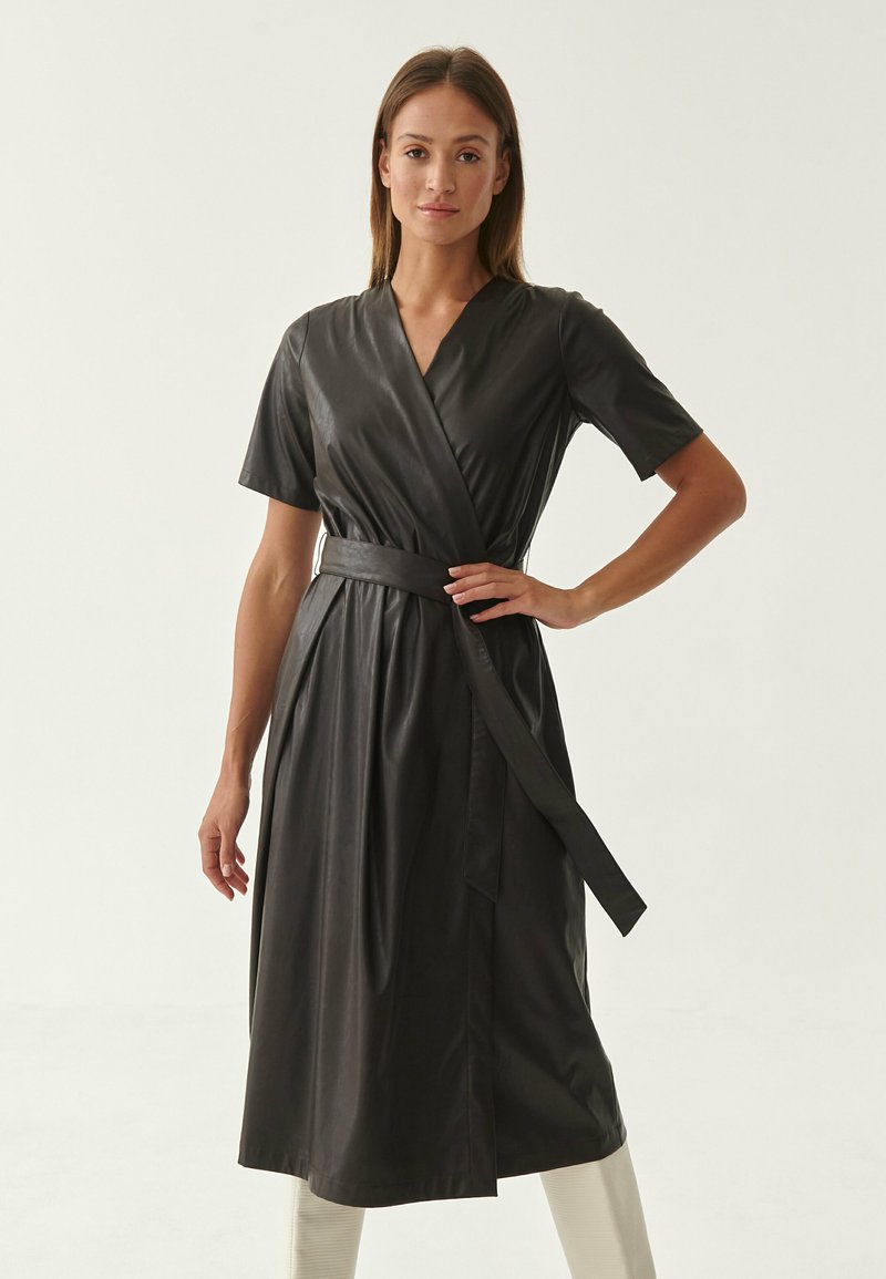Abito avvolgente in pelle nera con maniche corte, scollatura a V, cintura in vita e gonna fluida. Texture liscia, lunghezza al ginocchio, design minimalista.