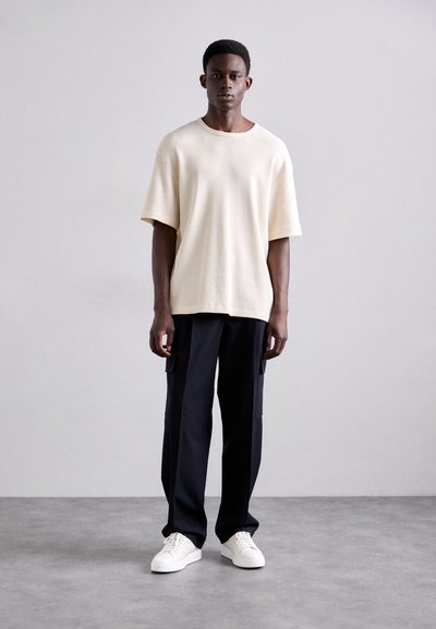 Camiseta de algodón texturizada color crema con mangas cortas, combinada con pantalones cargo oscuros con bolsillos laterales y zapatillas blancas.