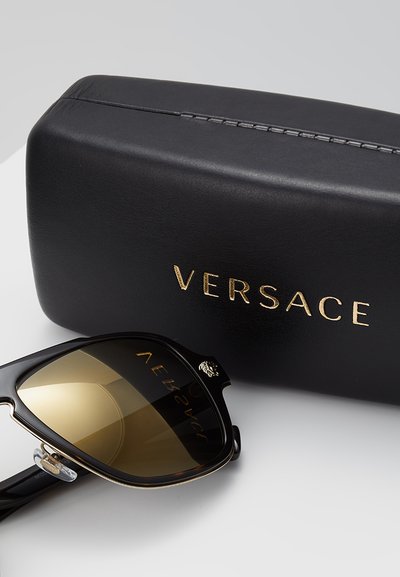 Svarte solbriller med gullfargede linser, med et dristig, vinklet design og logo på stengene, ved siden av et svart etui preget med "VERSACE."