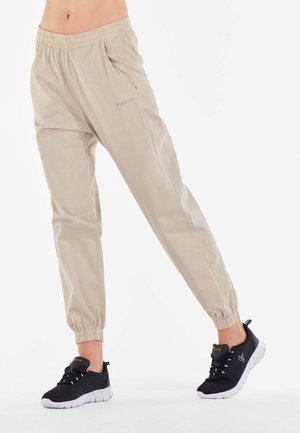 Beige Sweatpants mit einem elastischen Bund, gekrempelten Knöcheln und seitlichen Taschen. Kombiniert mit schwarzen Sneakers mit weißen Sohlen und Schnürsenkeln.