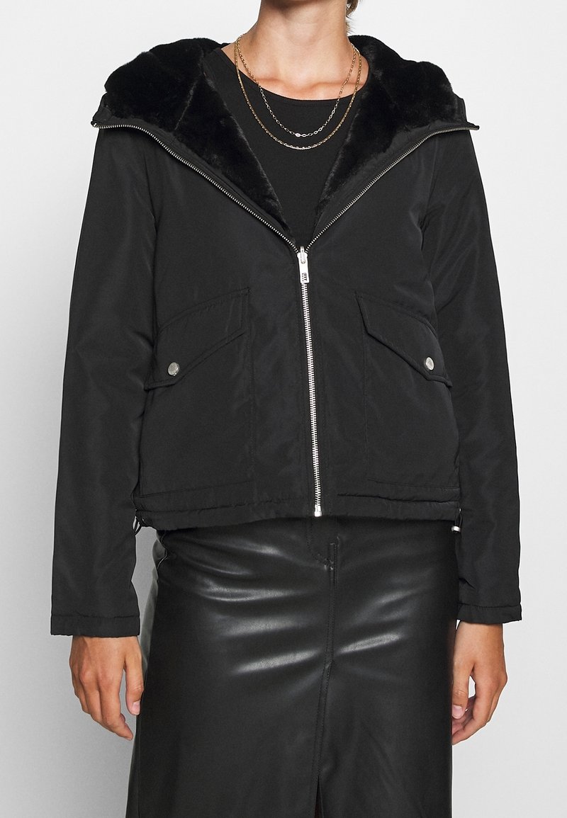 Veste noire zippée avec col en fausse fourrure et poches à rabat portée sur un top noir et une jupe en cuir, accessoirisée avec deux colliers fins en chaîne.