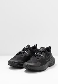 Zapatillas deportivas negras con parte superior de malla, suela de goma texturizada, cuello acolchado y acentos reflectantes. Diseñadas para la comodidad y el soporte.