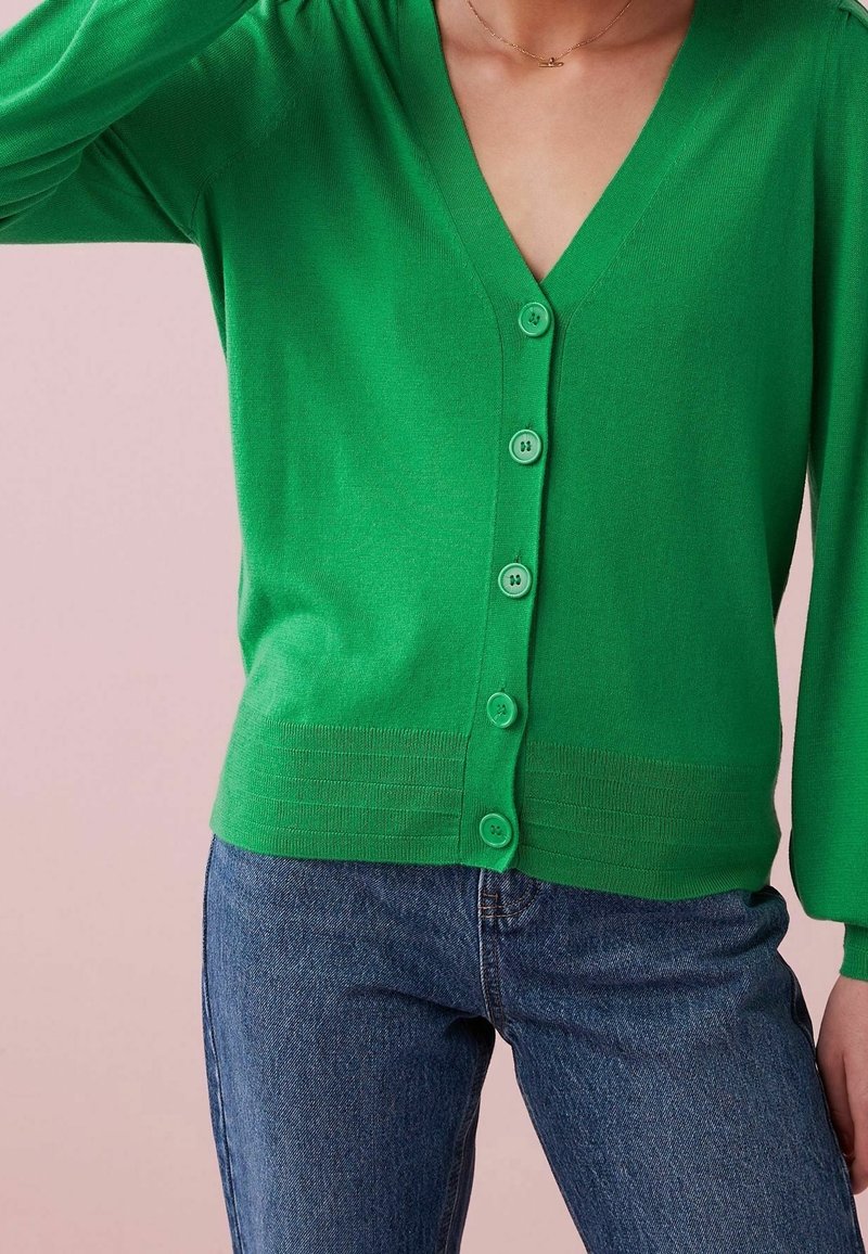 Cardigan verde a maglia con scollo a V, sei bottoni abbinati e orlo a coste. Abbinato a jeans blu, presentato su uno sfondo chiaro.