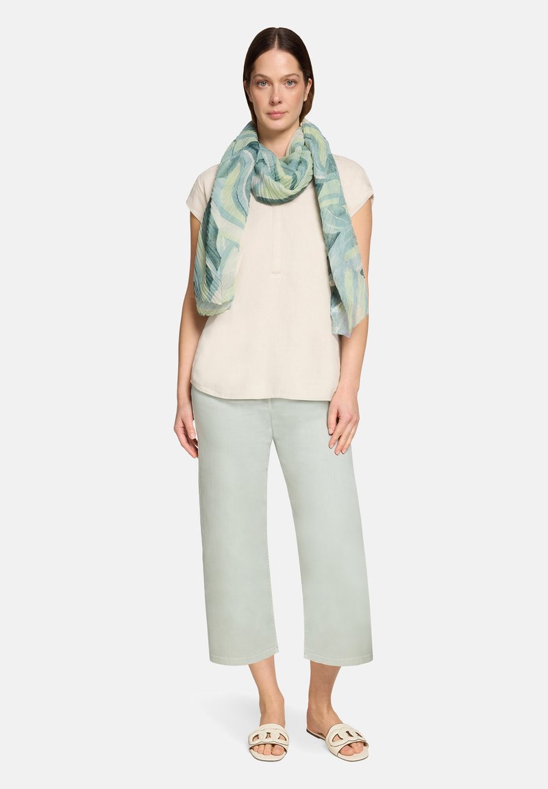 Vrouw in een lichtbeige top met korte mouwen, lichtgroene broek met wijde pijpen en cropped lengte, open teenslippers en een groen-blauw sjaaltje met patroon om haar nek.