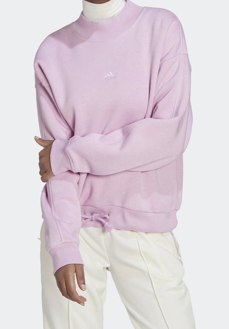 Personne portant un sweat Adidas violet clair avec un cordon à l'ourlet, par-dessus un col roulé blanc et un pantalon crème.