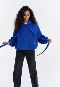 Hanorac albastru cu glugă, buzunar frontal, șnururi și un mic accent roz. Asortat cu pantaloni largi din denim închis la culoare.