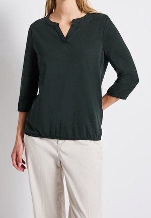 Femme portant un chemisier manches longues vert foncé avec un col en V et un pantalon beige clair sur un fond uni.