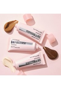 Tre tubi di Maybelline Perfectors con imballaggio rosa chiaro, accompagnati da campioni di trucco in beige cremoso, abbronzatura chiara e marrone scuro.