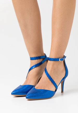 Zapatos de tacón alto azules con punta afilada y correas en el tobillo con bordes ondulados. Hechos de un material brillante con una superficie texturizada. Tacón de aguja.