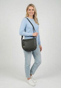 Mujer con suéter azul claro, jeans y zapatillas blancas, de pie con un bolso cruzado de cuero negro sobre un fondo gris.