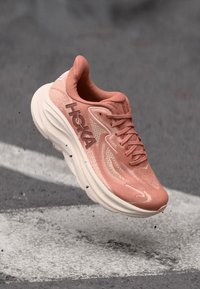 Baskets de course rose clair avec un dessus en maille, épaisse semelle intermédiaire blanche et détails texturés. Présente un logo "HOKA" bien visible sur le côté.