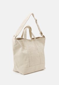 Borsa a spalla in tela beige con due manici superiori e una tracolla regolabile. Presenta cuciture verticali e una base strutturata.