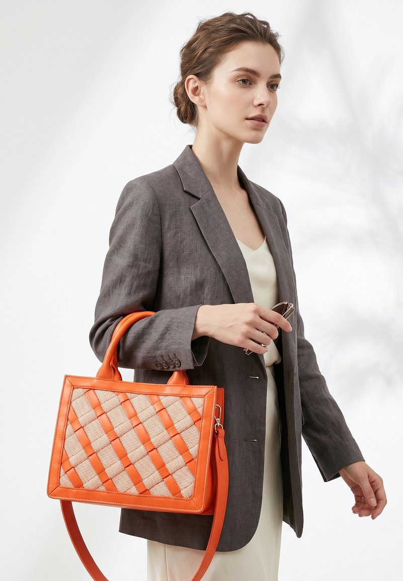 Femme en blazer gris tenant un sac à main tissé orange et une paire de lunettes sur un fond clair neutre.