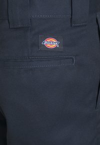 Ciemnoniebieskie spodnie z wytrzymałego materiału, wyposażone w tylną kieszeń oraz prostokątną naszywkę marki Dickies w czerwono-żółtych akcentach.