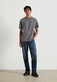 Graues T-Shirt mit Rundhalsausschnitt aus weichem Stoff, kombiniert mit blauen Jeans. Das Modell trägt schwarze Schuhe mit strukturierten Details. Einfaches, lässiges Outfit.