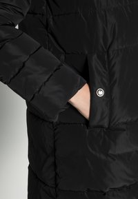 Schwarze Daunenjacke mit gestepptem Muster, ausgestattet mit einer Seitentasche und einem sichtbaren Druckknopf. Der Stoff hat eine glatte, glänzende Textur.