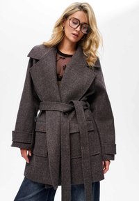 Bruine houndstooth wollen blendjas met een oversized kraag, een ceintuur in de taille, voorzakken en brede mouwen. Draagt over een transparante bloemen top.