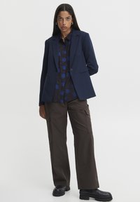 Blazer marine à la coupe structurée et à un bouton, porté sur une chemise à motifs bleus et noirs ; associé à un pantalon cargo marron et des bottes noires.