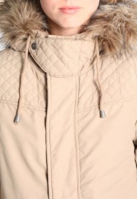 Giubbotto trapuntato beige con cappuccio in pelliccia sintetica, dotato di lacci regolabili e accessori in metallo argentato. Il tessuto è liscio e leggero.