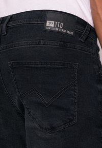 Los jeans de mezclilla negra cuentan con un bolsillo trasero distintivo con un diseño de costura en forma de V. La etiqueta dice "TTD Tom Tailor Denim Brand."