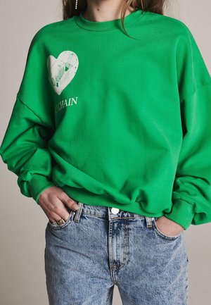 Personne portant un sweat-shirt vert vif avec un cœur blanc et le texte « UNCHAIN », assorti à un jean en denim bleu clair, les mains dans les poches.